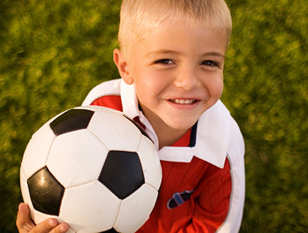ילד בלונדיני באדום על דשא מחזיק כדורגל ומחייך (צילום: jupiter images)