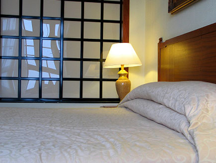 מיטה עם מצעים לבנים ולידה מנורת לילה וחלון משובץ (צילום: Naomi Hasegawa, Istock)