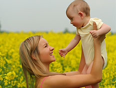 בחורה בורוד מרימה תינוק ומחייכת בשדה פרחים צהובים (צילום: אור גץ, jupiter images)