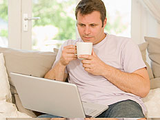 גבר בלבן יושב עם מחשב נישא עליו וספל בסלון מאחוריו (צילום: jupiter images)