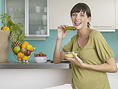 אישה בהריון עומדת במטבח, נינוחה מחייכת ואוכלת (צילום: jupiter images)