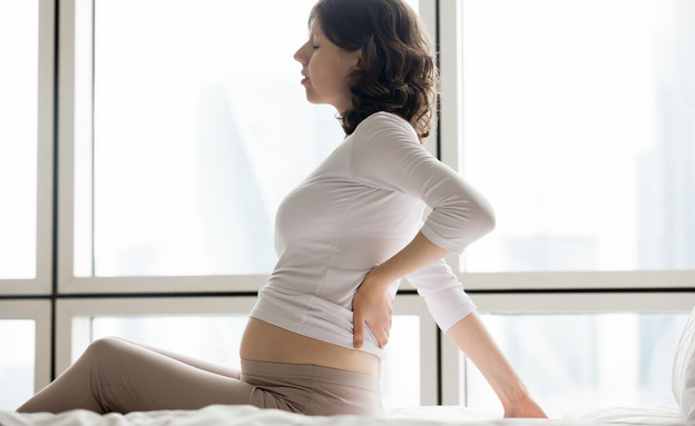 אישה בהיריון יושבת על מיטה ומחזיקה את הגב (אילוסטרציה: fizkes, shutterstock)
