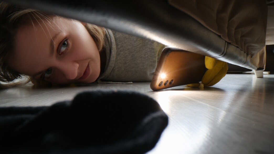 אישה בודקת מתחת למיטה עם פנס של הטלפון (צילום: H_Ko, SHUTTERSTOCK)