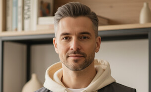 גבר עומד ליד מדף ספרים ומסתכל למצלמה (AI: mako)