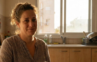 אישה יושבת במטבח ביתה ומסתכלת למצלמה (AI: mako)