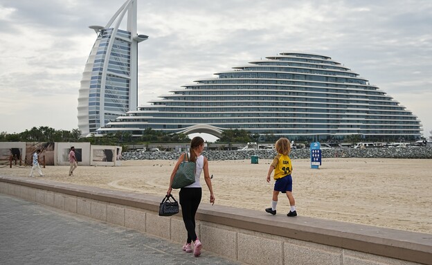 אישה וילדה צועדים לאורך חוף הים בדובאי, כשברקע מלונות 