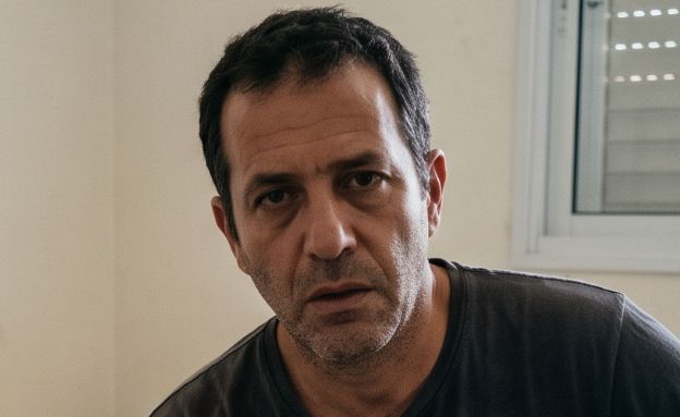 גבר יושב על מיטה עם אישה ברקע (AI: mako)
