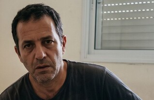 גבר יושב על מיטה עם אישה ברקע (AI: mako)